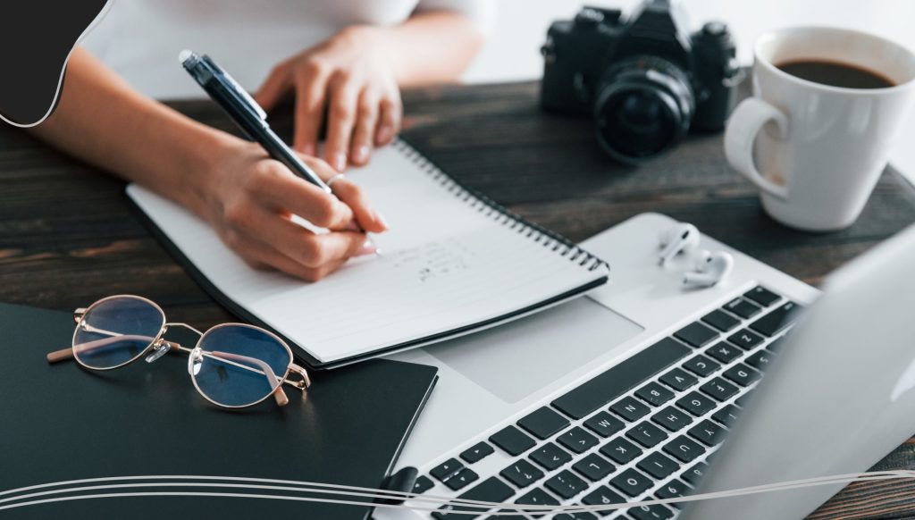 Person writing in a notebook on a desk with a camera and laptop