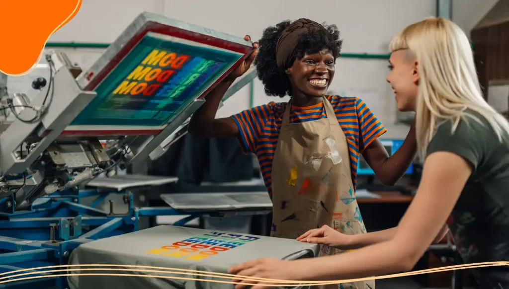 Smiling interracial print shop female workers silkscreen printing t-shirt.